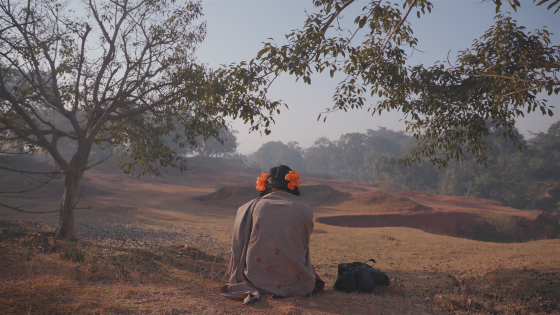 A girl with orange bows sits with her back on the sand next to a tree